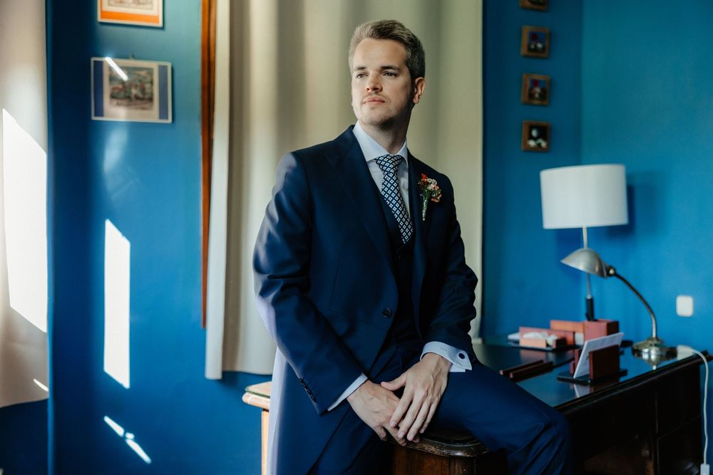 Business professional in a navy suit stands in a modern office space with bright blue walls and natural lighting.