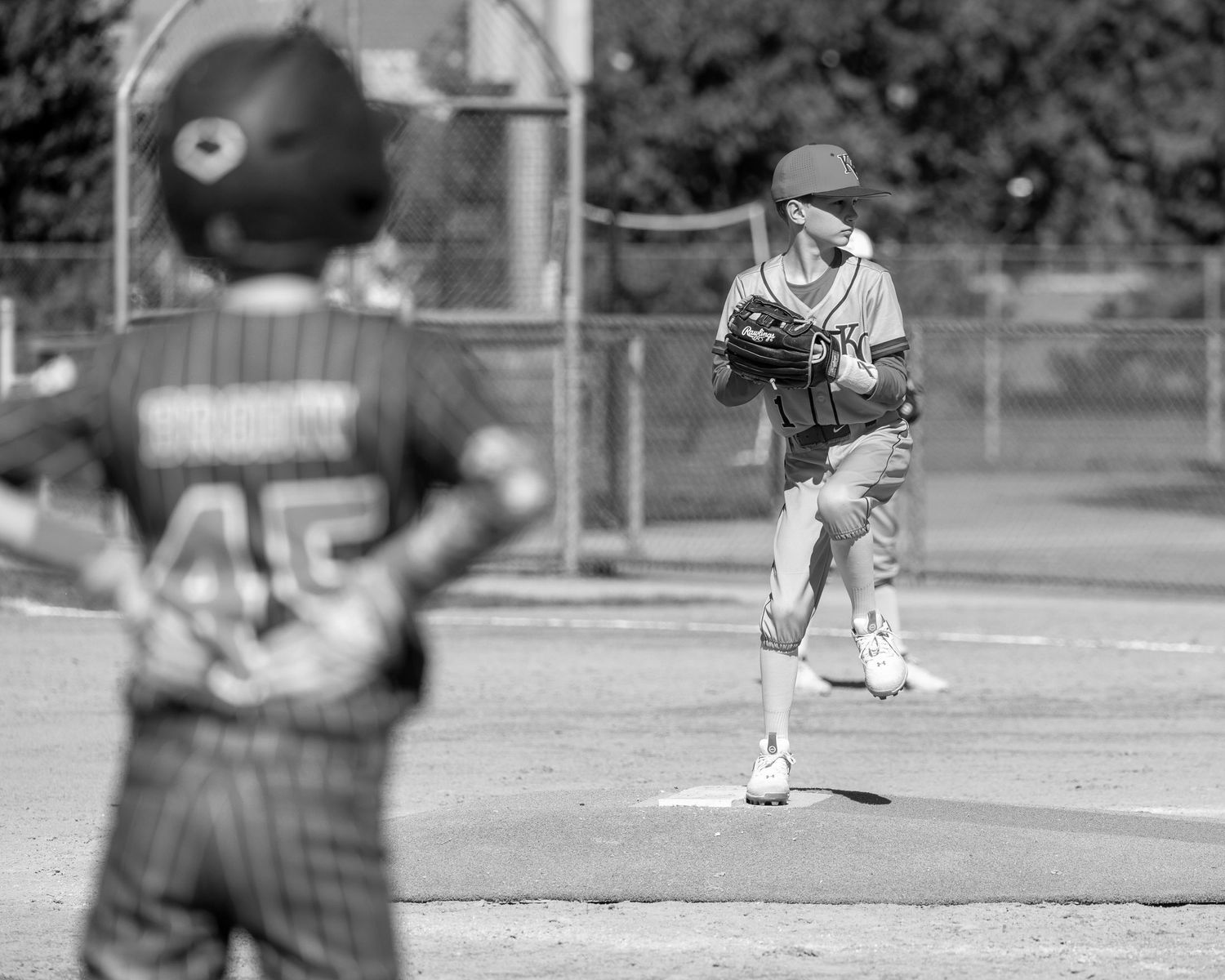 Black and white baseball action shot showing a pitcher in mid-delivery on the mound during a youth game.