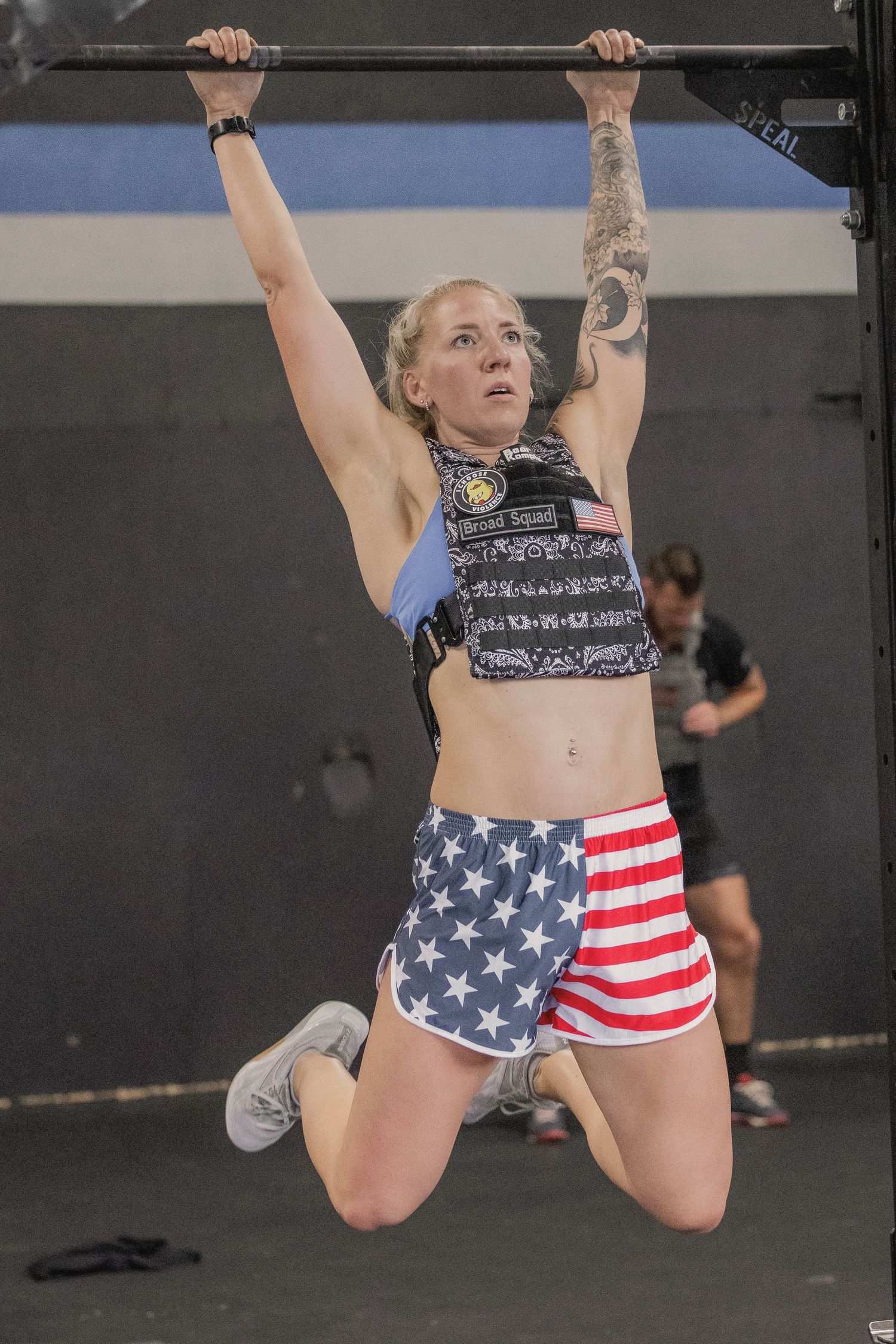 Athlete doing pull-ups wearing American flag shorts and tactical vest in gym setting.