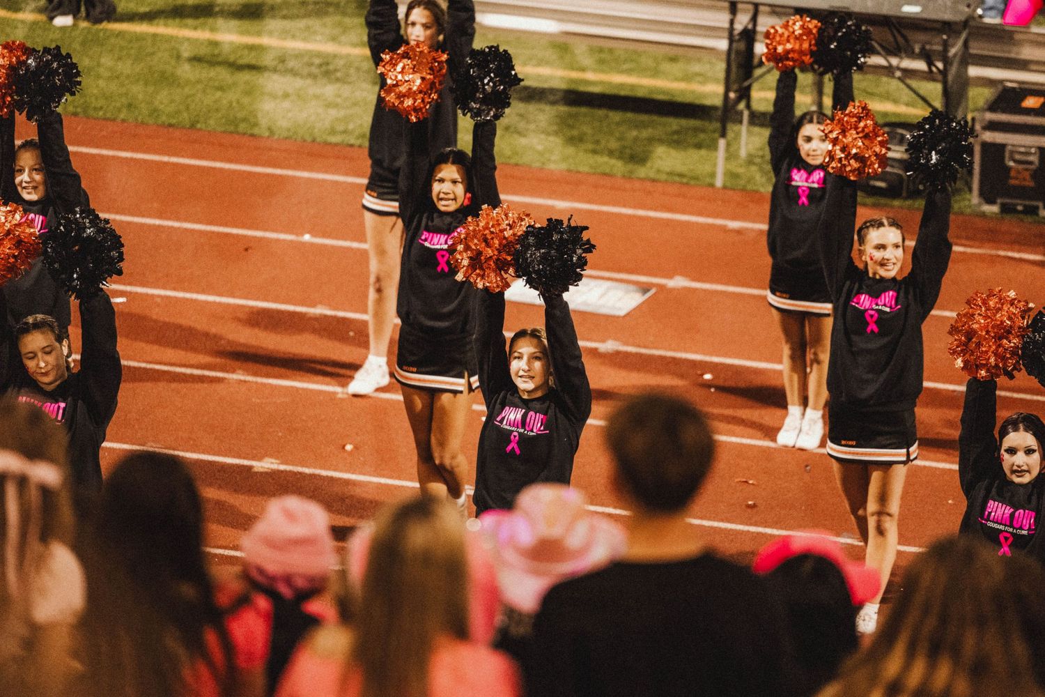 Cheerleaders in black uniforms perform a synchronized routine with orange pom poms on a track field.