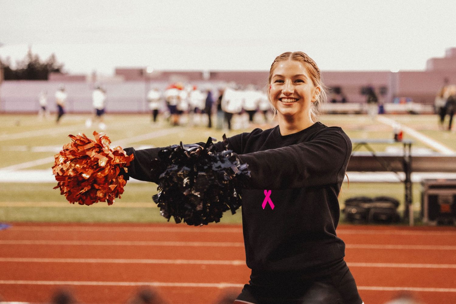 A cheerleader in black uniform performs with pom poms on an outdoor track.