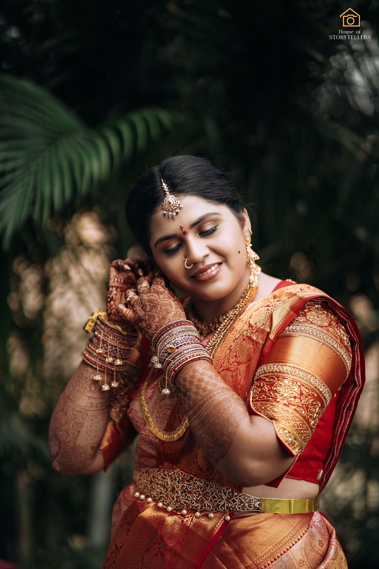 Bride in orange silk saree with traditional henna and jewelry poses near palm leaves.