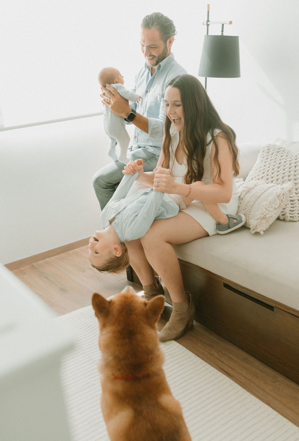 Family relaxing on couch with baby while dog watches from below.