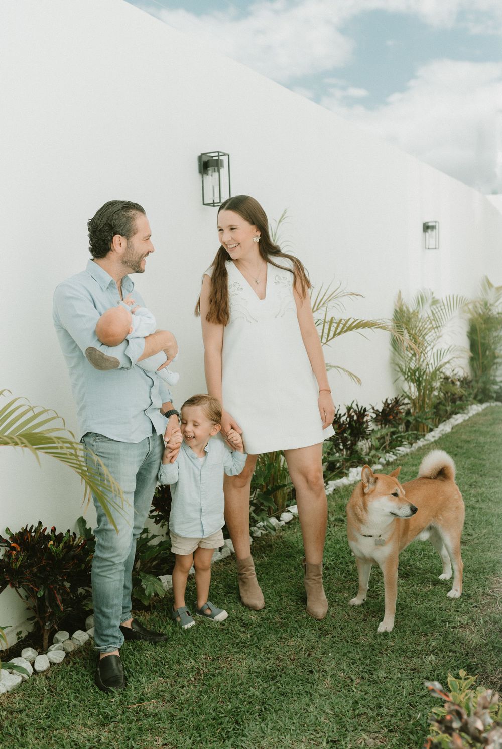 Family standing together with their Shiba Inu dog against white wall outdoors.
