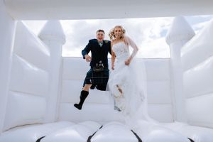 Playful couple jumping together on a white bouncy castle in wedding attire.