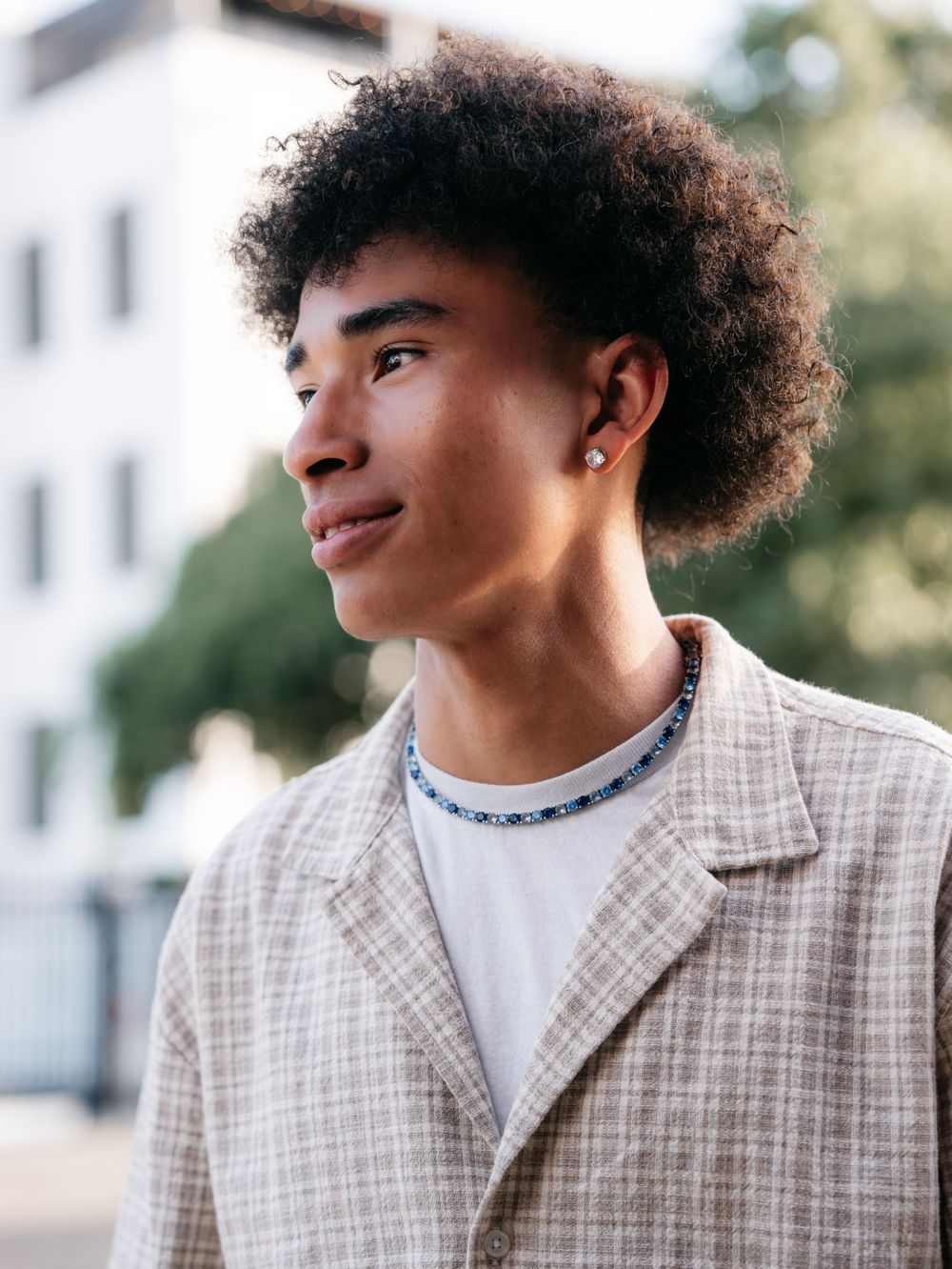 Young man with curly hair wearing a plaid blazer and beaded necklace looking away outdoors.