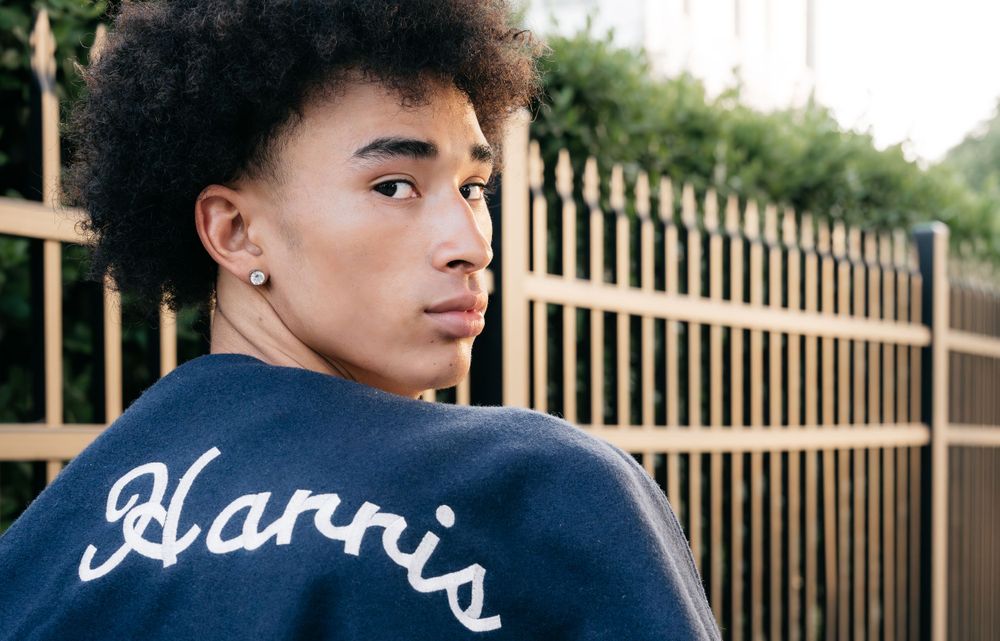 Young man with curly hair wearing a Harris varsity jacket looking back over his shoulder outdoors.