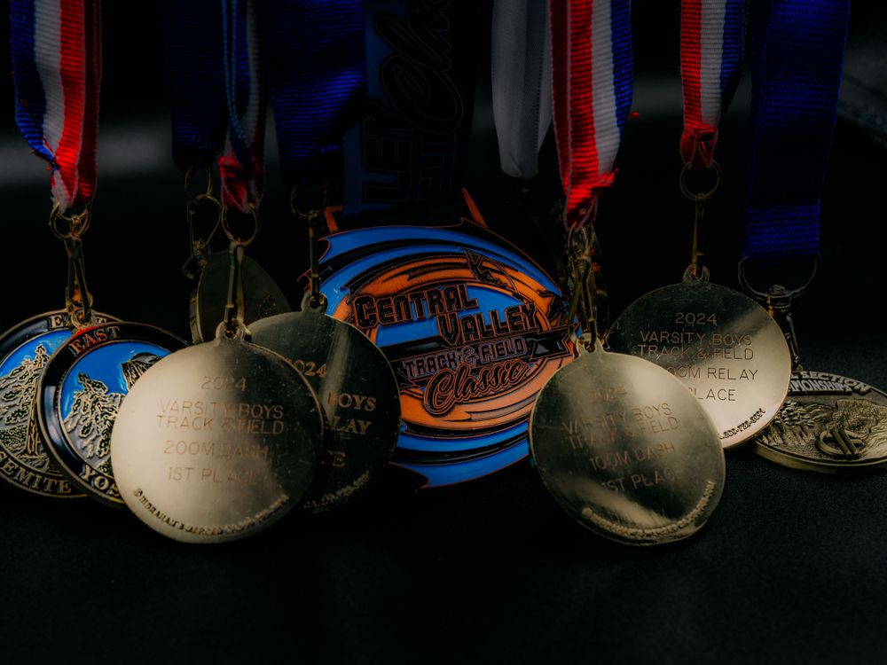Collection of gold and silver sports medals with red white and blue ribbons on a dark surface.