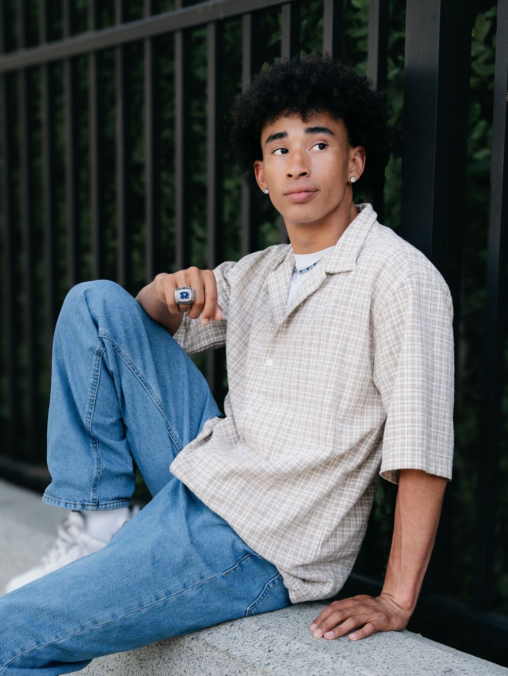 Young man in plaid shirt and jeans sitting on concrete ledge outdoors.
