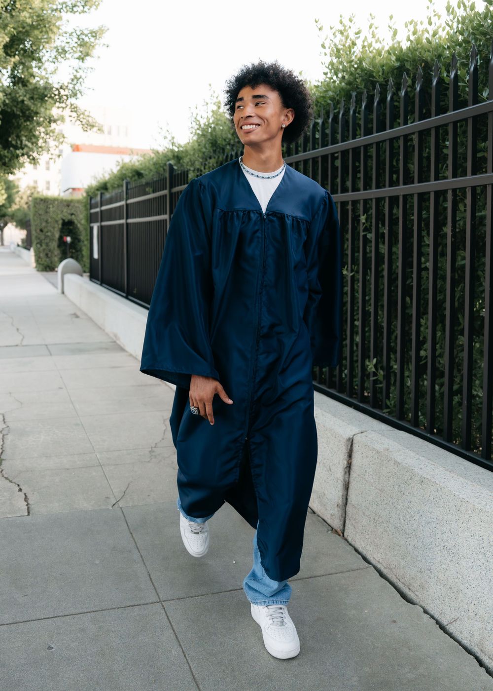 Smiling graduate in navy cap and gown walking on sidewalk outdoors.