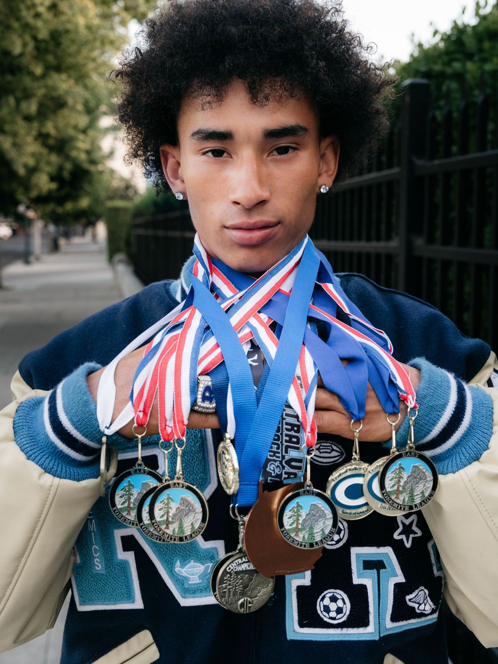 Young male athlete proudly displaying multiple sports medals around his neck wearing a varsity jacket.