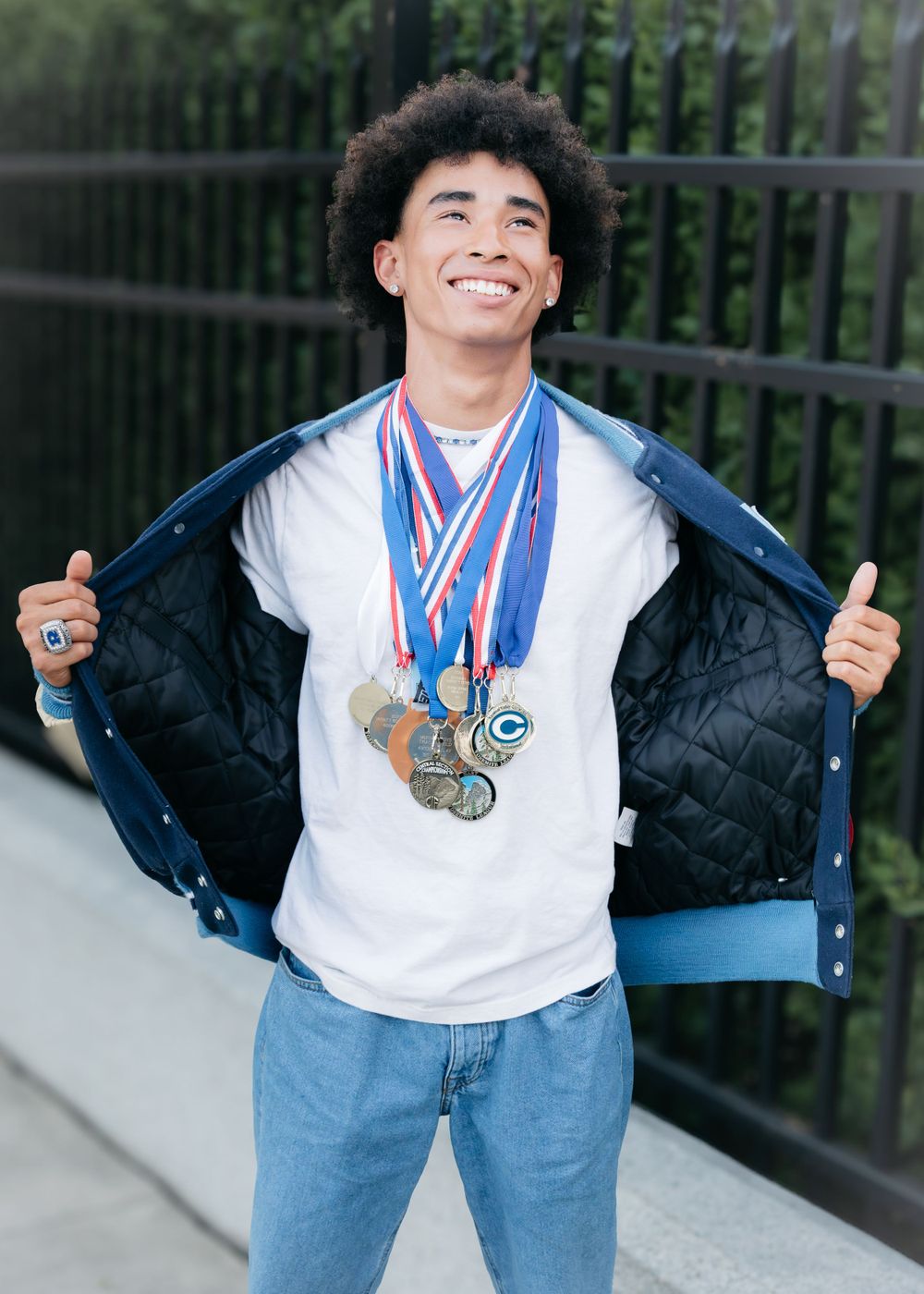 Smiling young man wearing multiple sports medals opening his blue jacket outdoors near a fence.