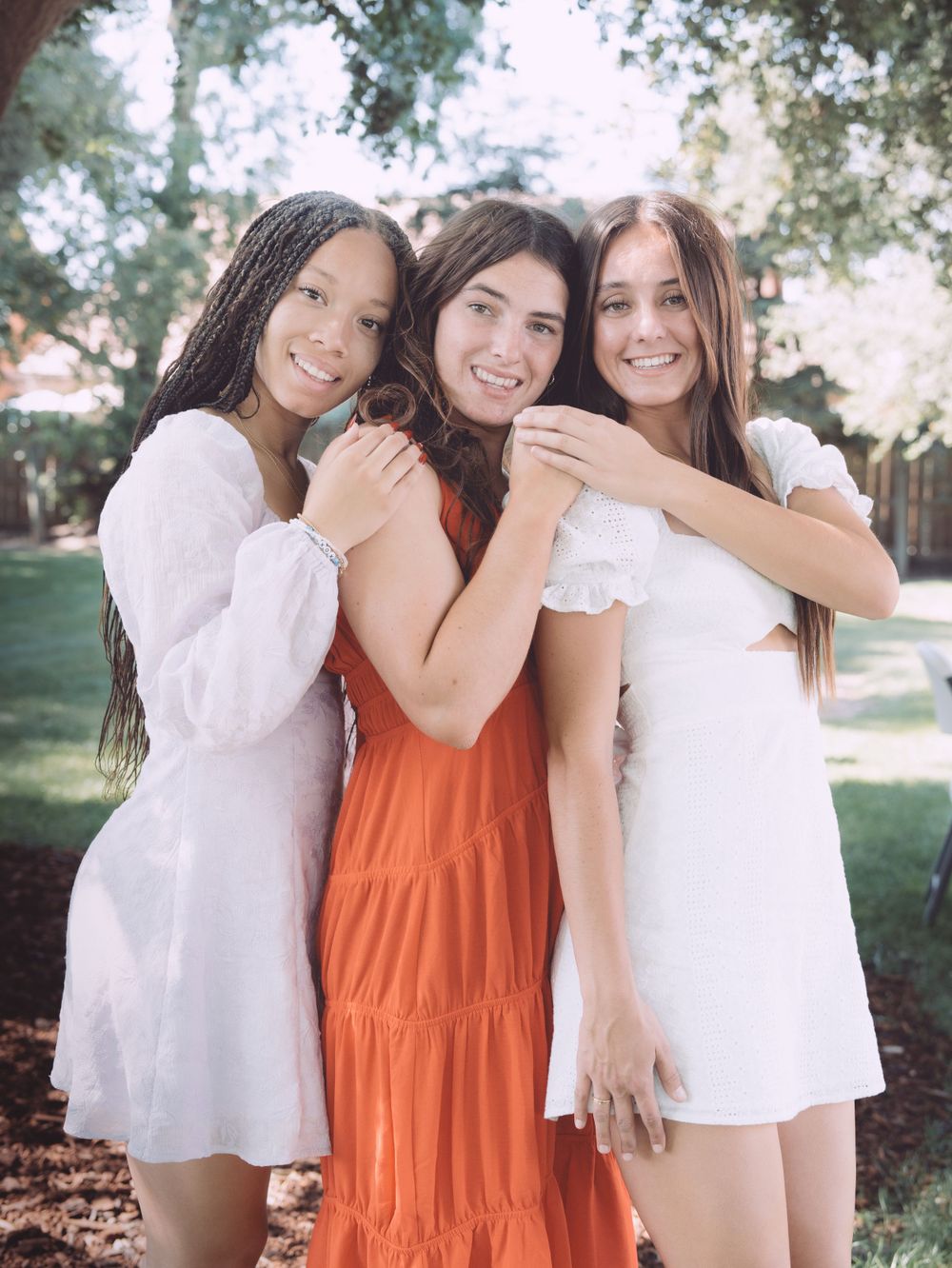 Three young women in white and orange dresses smile and embrace outdoors in a sunny garden setting.