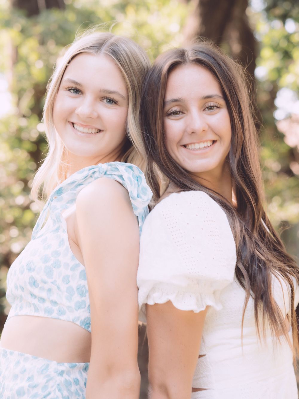 Two smiling young women posing back to back outdoors in summer tops.
