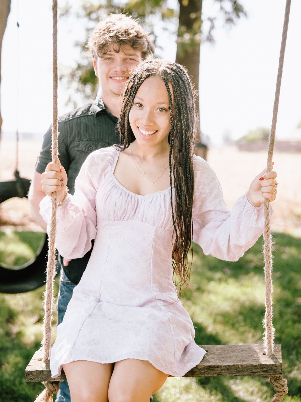 Young woman in pink dress sitting on wooden swing with young man behind her.