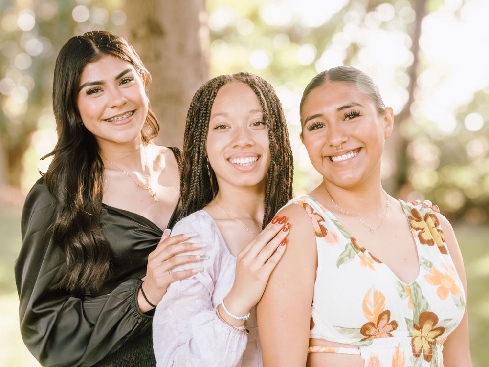 Three young women smiling together outdoors in summer outfits.
