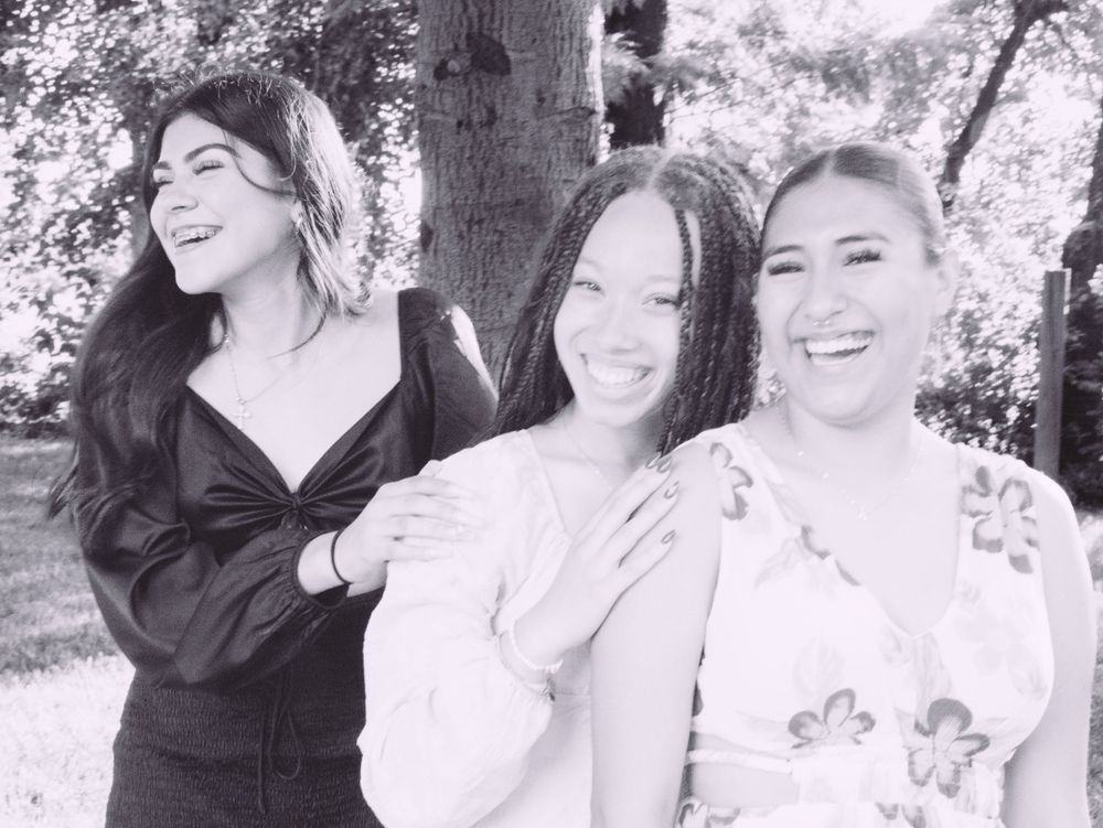 Three smiling women pose together outdoors near trees in this black and white photo.