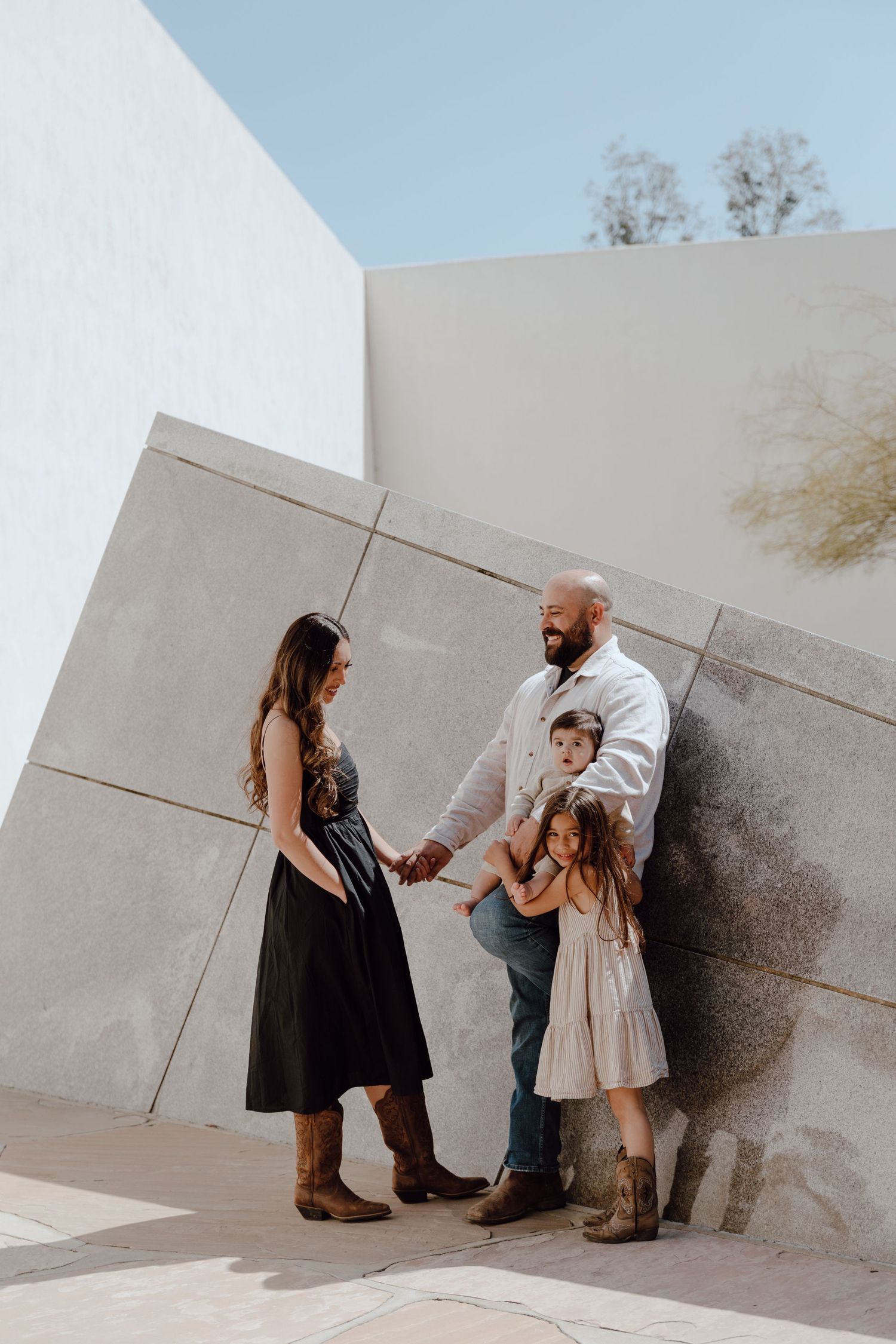 Family of four in casual attire standing against modern white wall architecture.