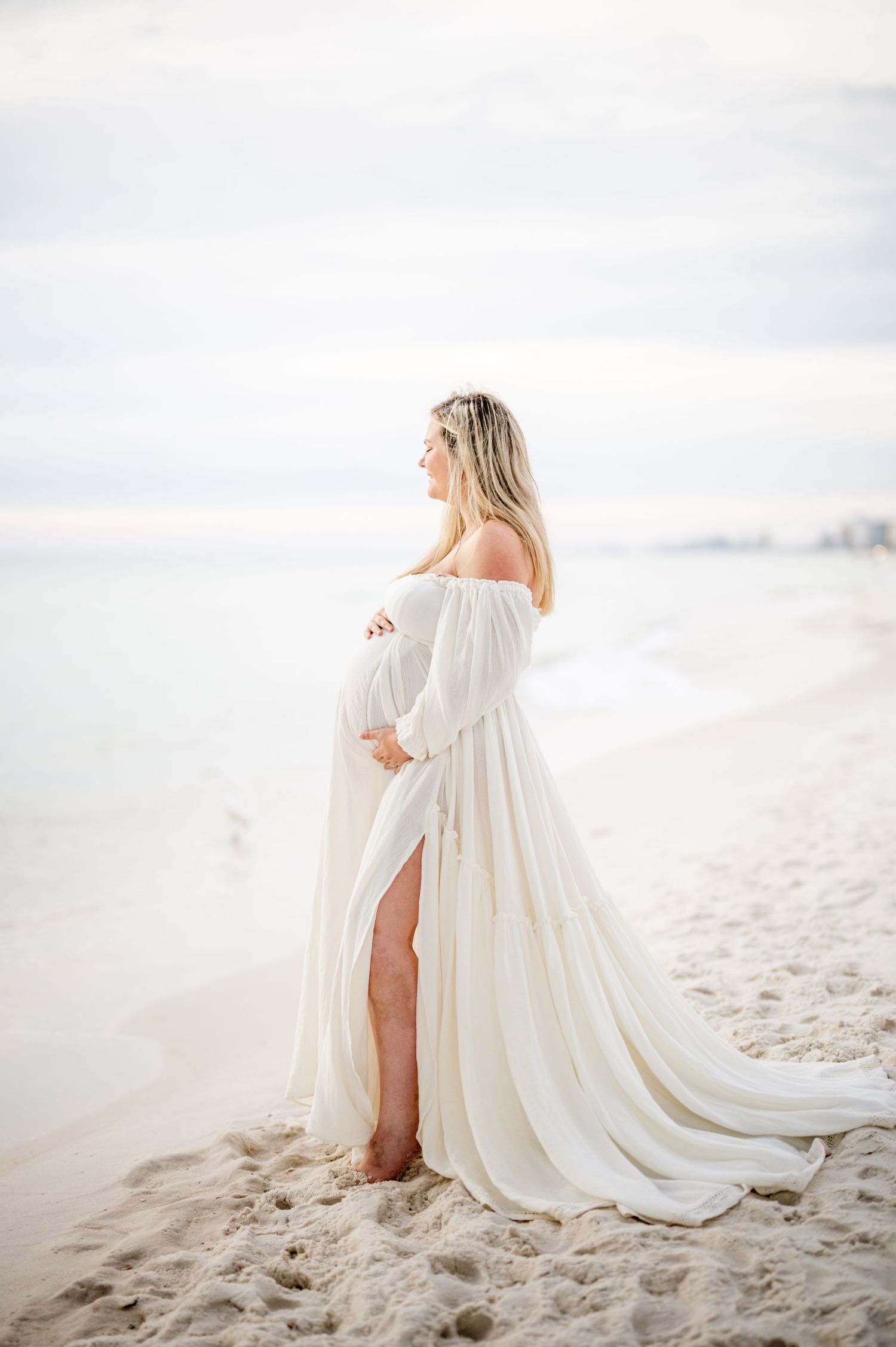 Maternity beach portrait in an ethereal white flowing gown against ocean backdrop.