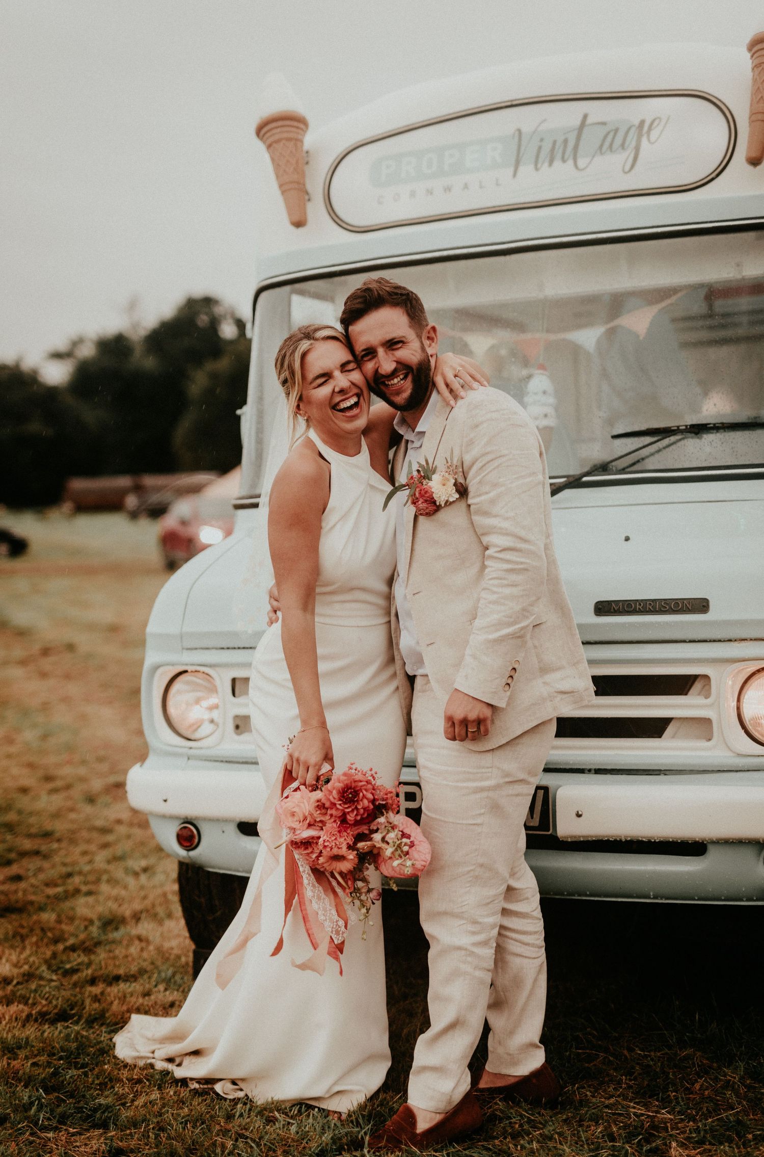 A romantic wedding couple poses together near a vintage white vehicle in a rustic outdoor setting at sunset.