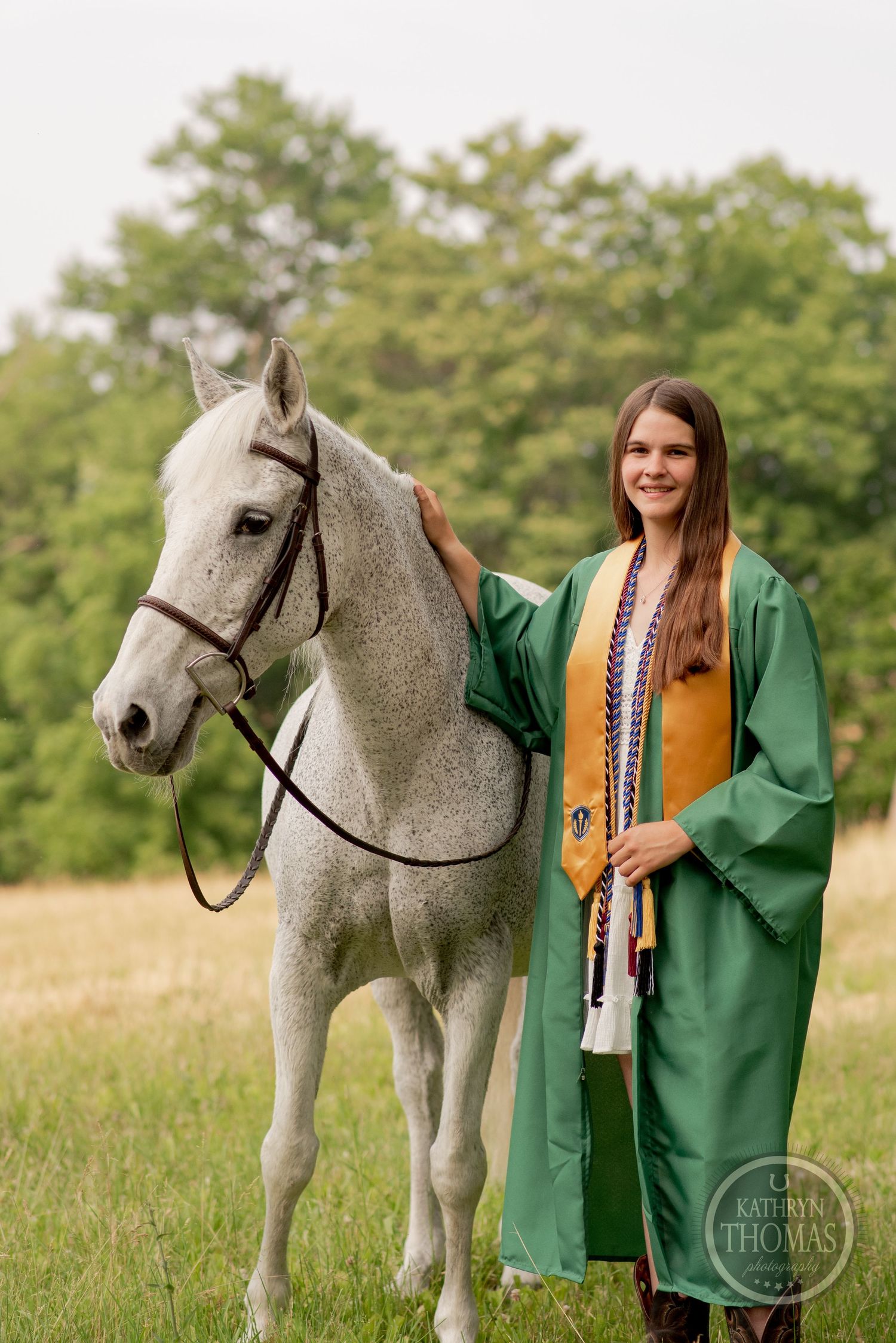 A graduate in green robes poses with a beautiful gray horse in a sunny meadow.