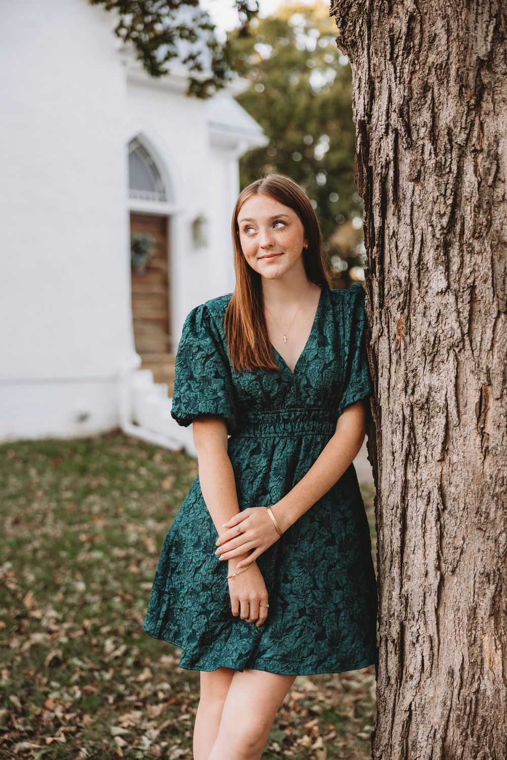 senior girl fall session in front of little white chapel leaning on a tree. Christmas green dress red hair blue eyes