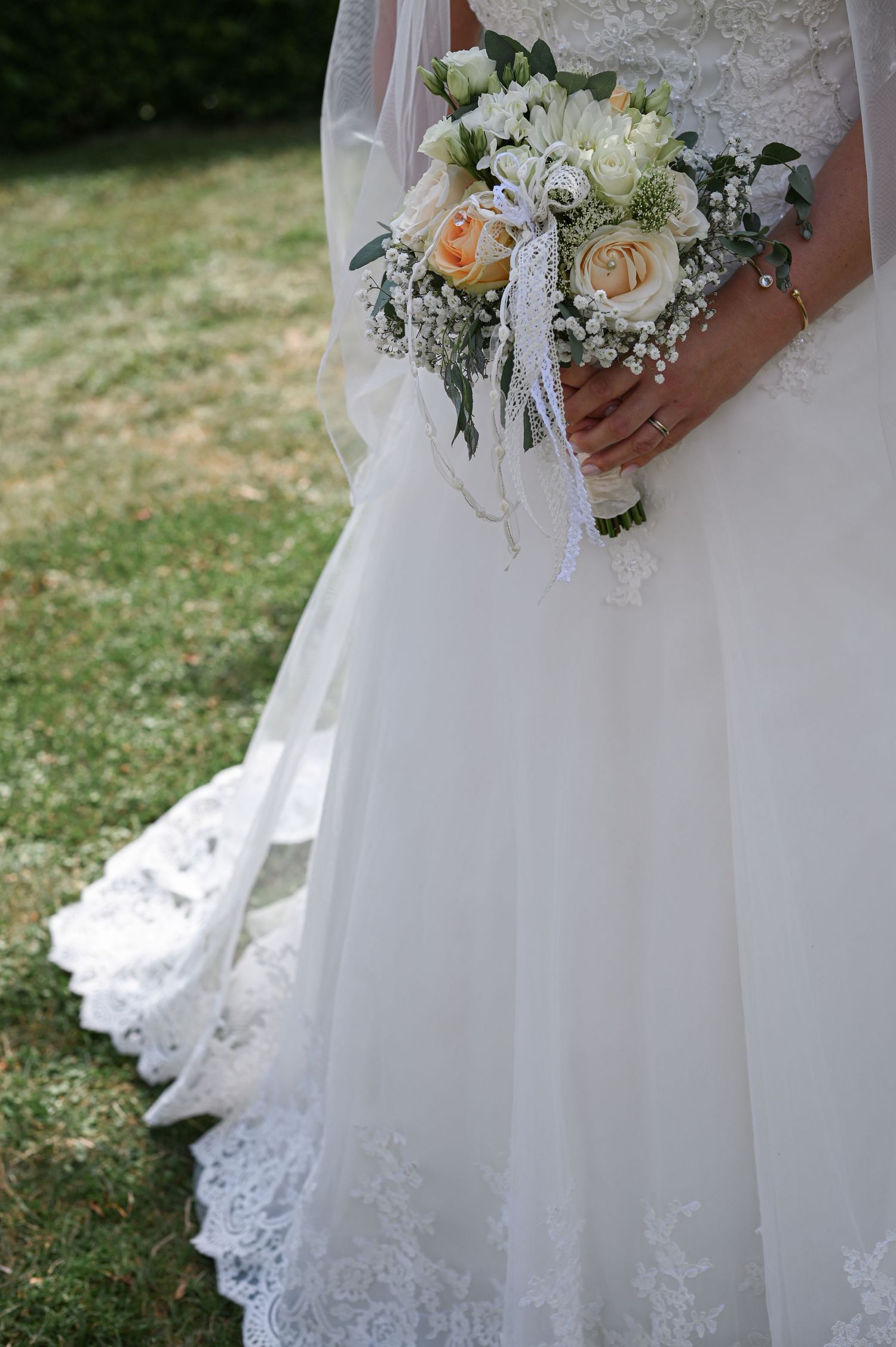 Ein Brautstrauß aus pfirsichfarbenen und weißen Rosen kontrastiert mit einem Hochzeitskleid aus weißer Spitze auf einer Wiese im Freien.