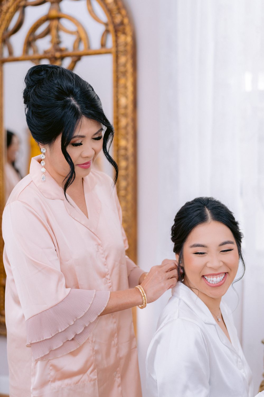 Two people sharing a joyful moment while getting ready in matching pink and white robes with an ornate mirror in background.
