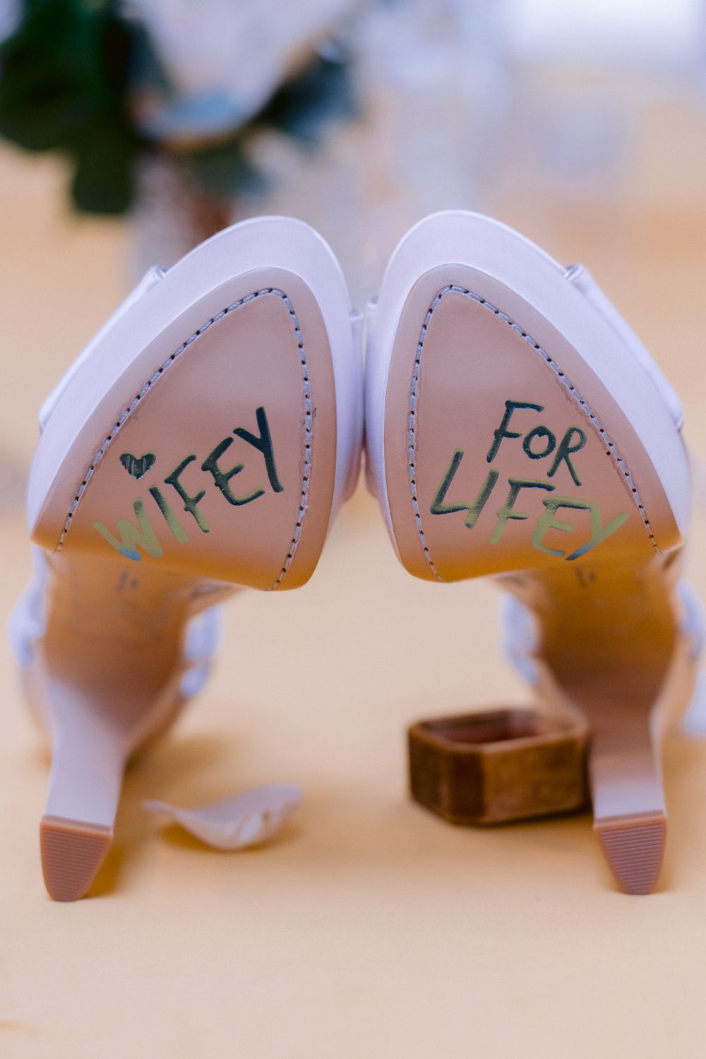 A pair of white high-heeled shoes with 'Wifey for Life' written on the soles, positioned upside down on a surface.