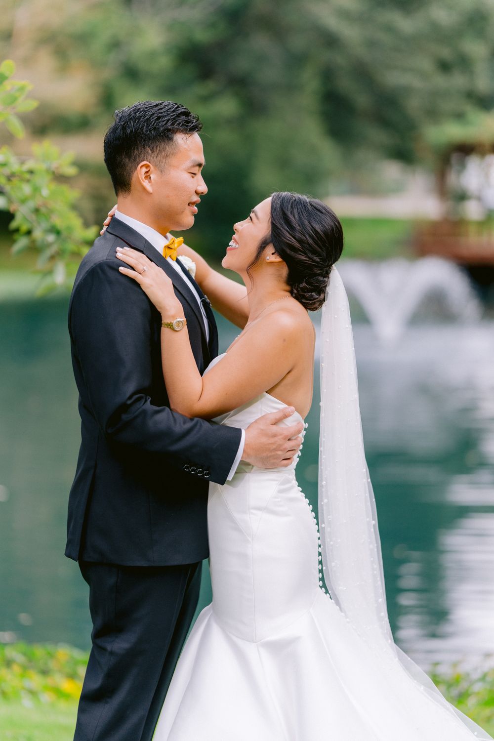Newlywed couple embraces outdoors by scenic pond with fountain during romantic wedding photo session.
