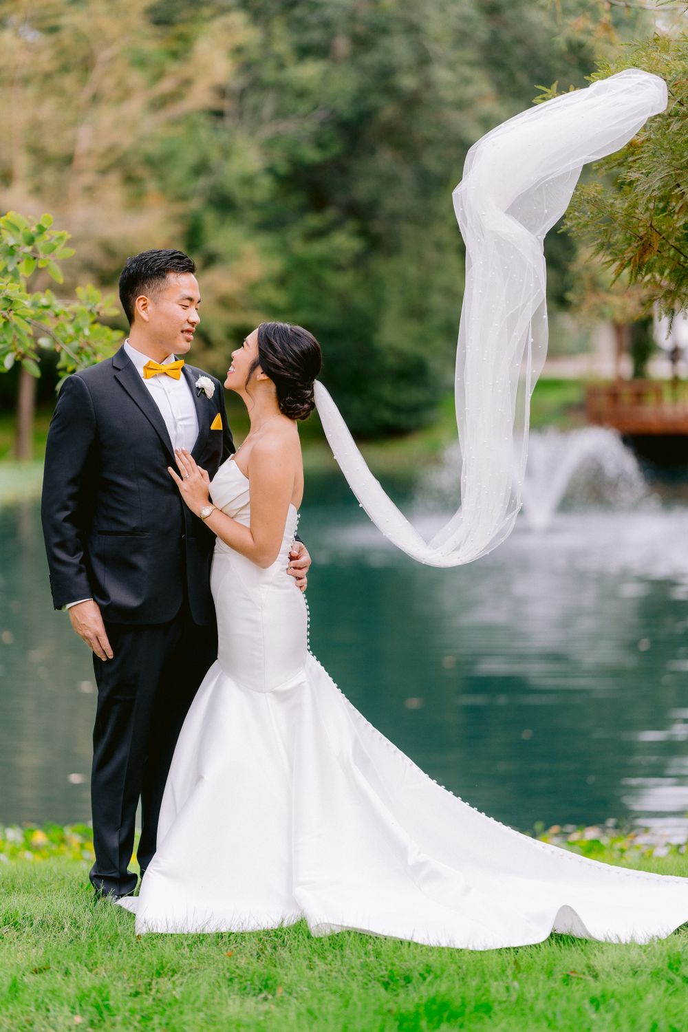 A newlywed couple poses by a lake while the bride's long veil gracefully flows in the wind.