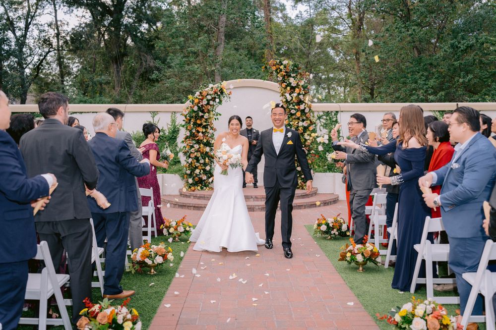A couple walks down an outdoor aisle decorated with sunflowers and yellow floral arch while guests celebrate at the ceremony.