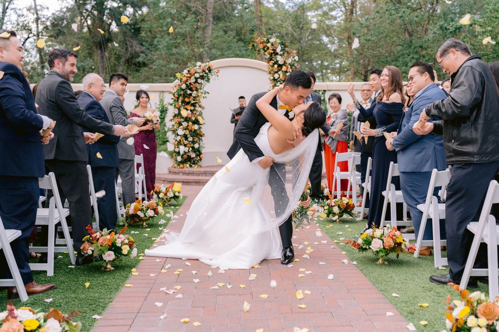 A couple embraces and kisses on a floral-lined outdoor aisle surrounded by seated guests at a wedding ceremony.