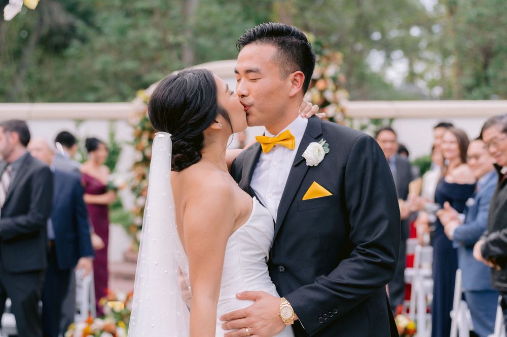 A bride and groom share their first kiss at an outdoor wedding ceremony while guests look on in the background.
