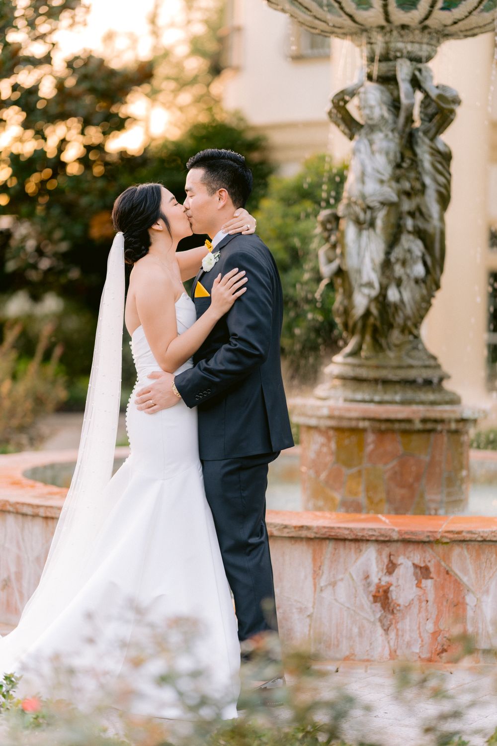 A romantic wedding moment captured in front of an ornate stone fountain with blurred bokeh sunlight in the background.