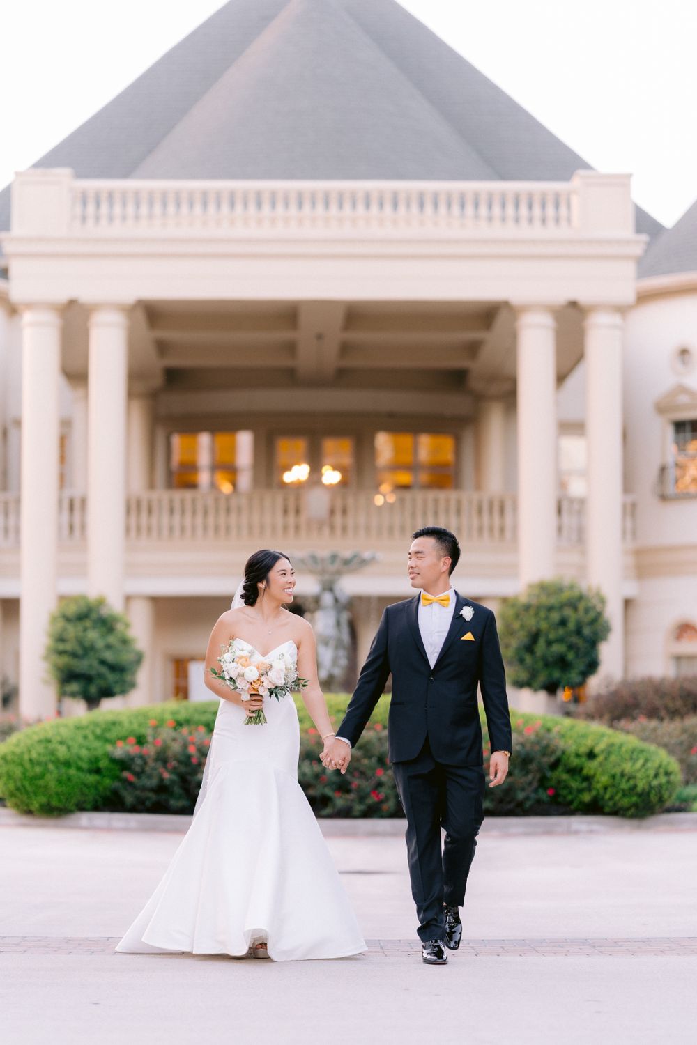 A couple walks hand in hand outside an elegant white mansion with columns and warm interior lighting at dusk.