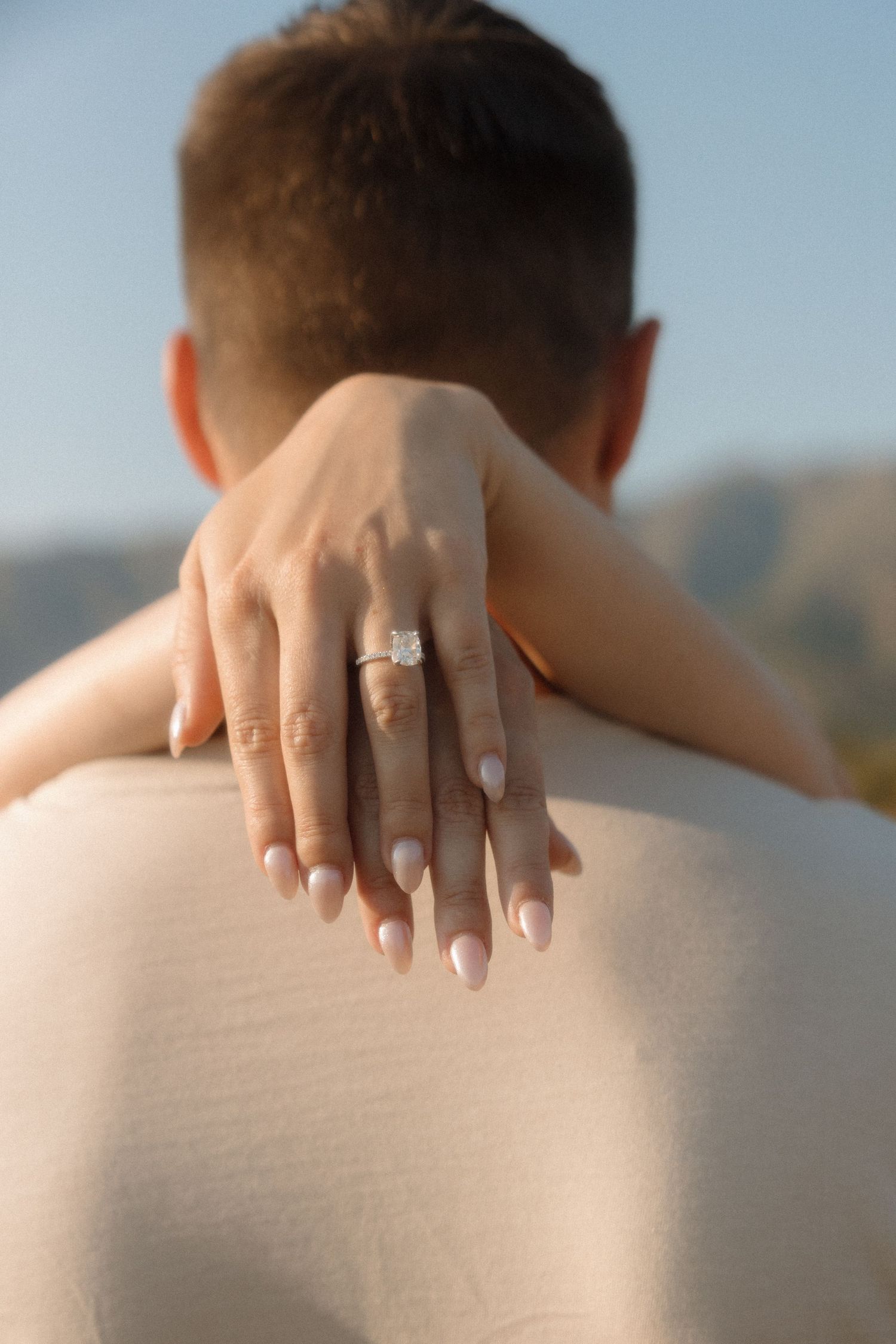 Close-up shot of hands embracing with an engagement ring displayed against a soft outdoor background.