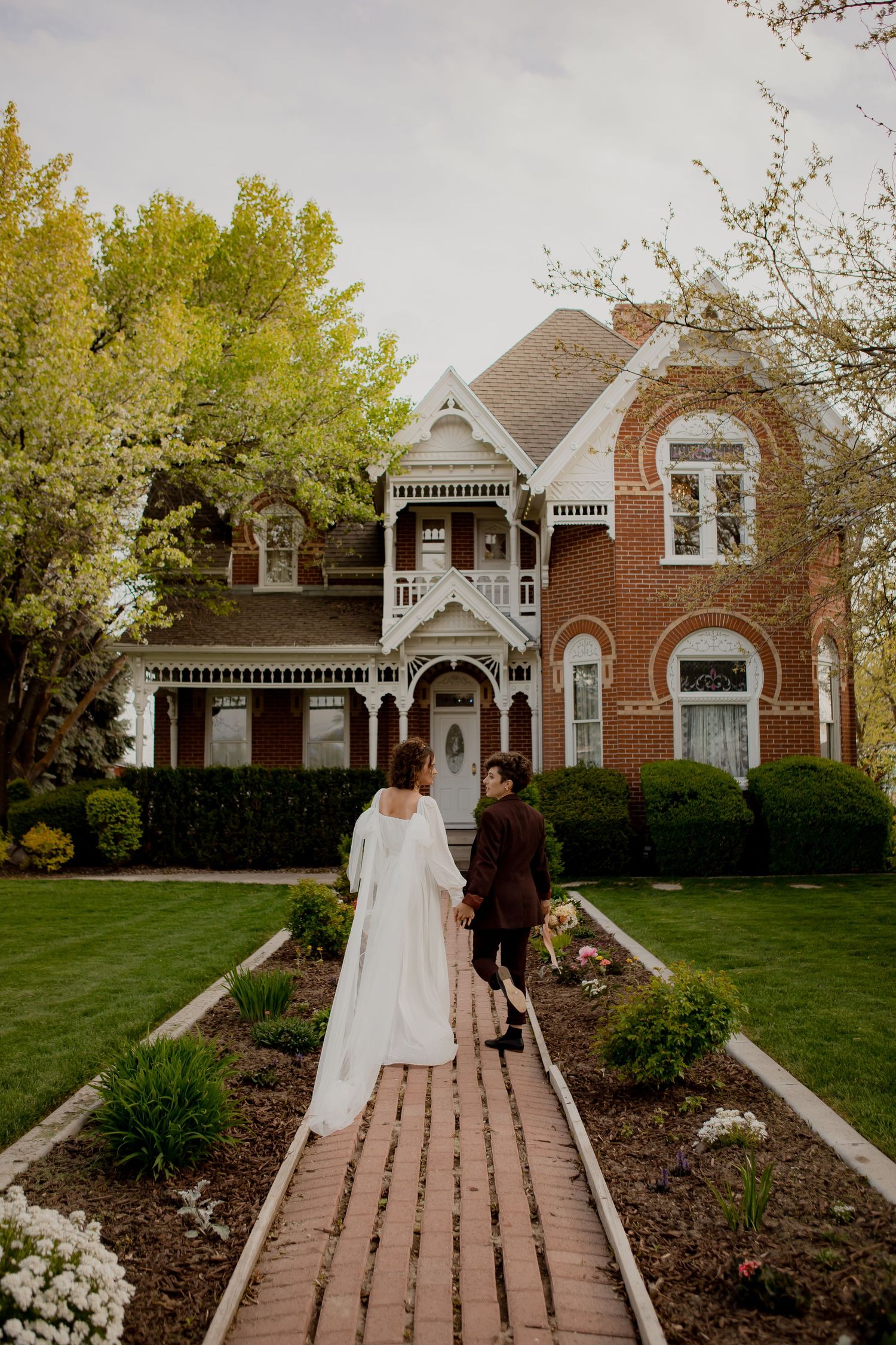 Couple walking path to beautiful Victorian brick mansion with white trim and landscaped garden.