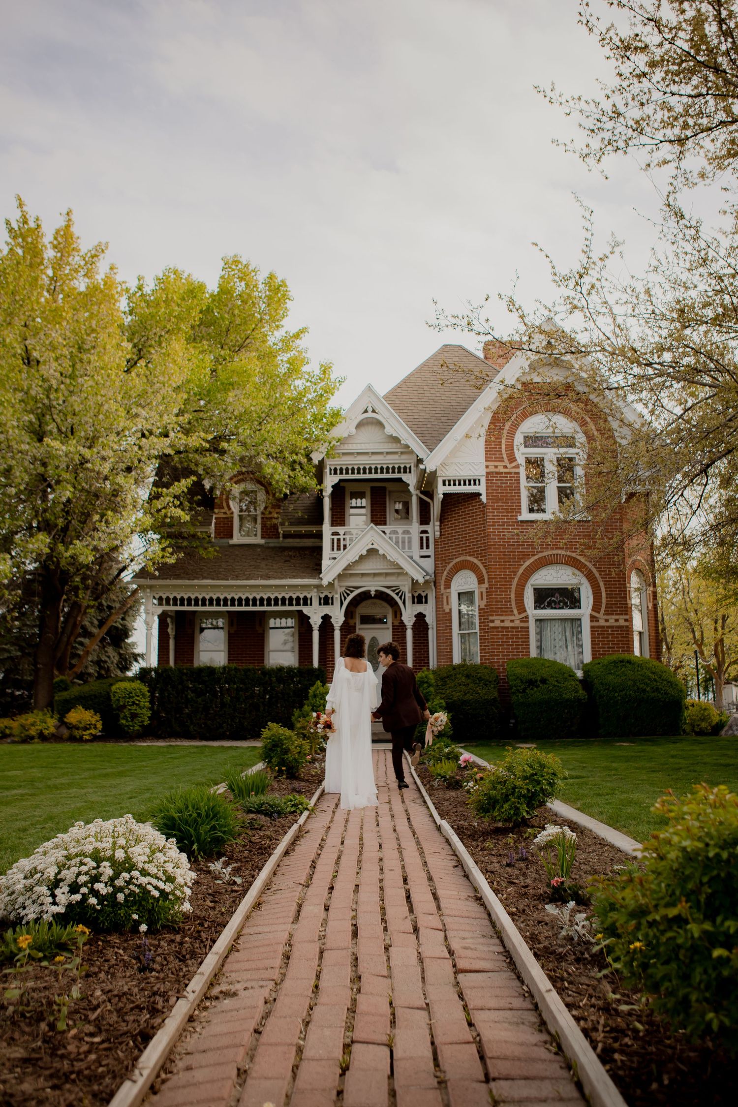 Wedding couple approaches elegant Victorian home along brick pathway framed by spring gardens.