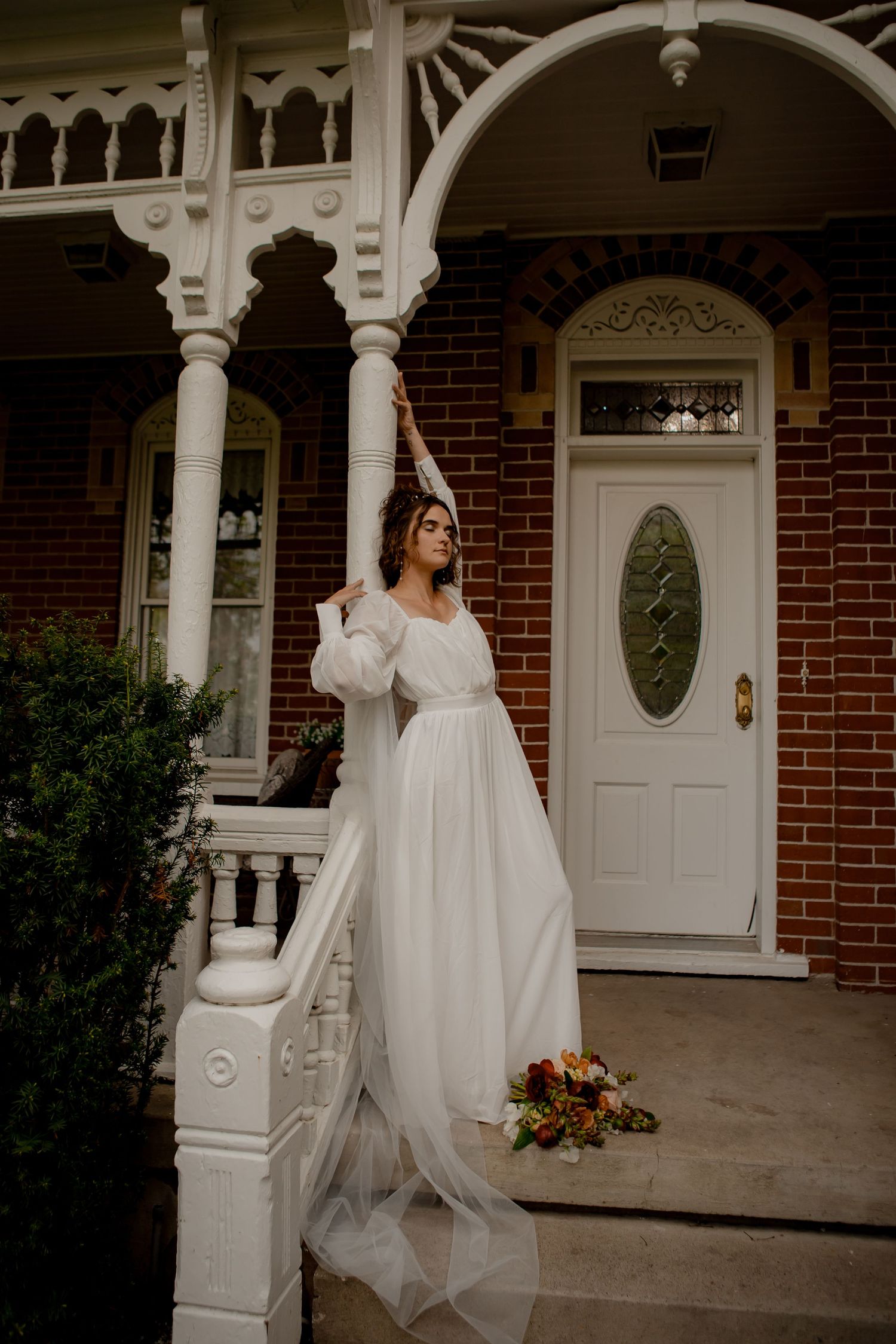 Bride in flowing white dress poses elegantly on Victorian porch steps.