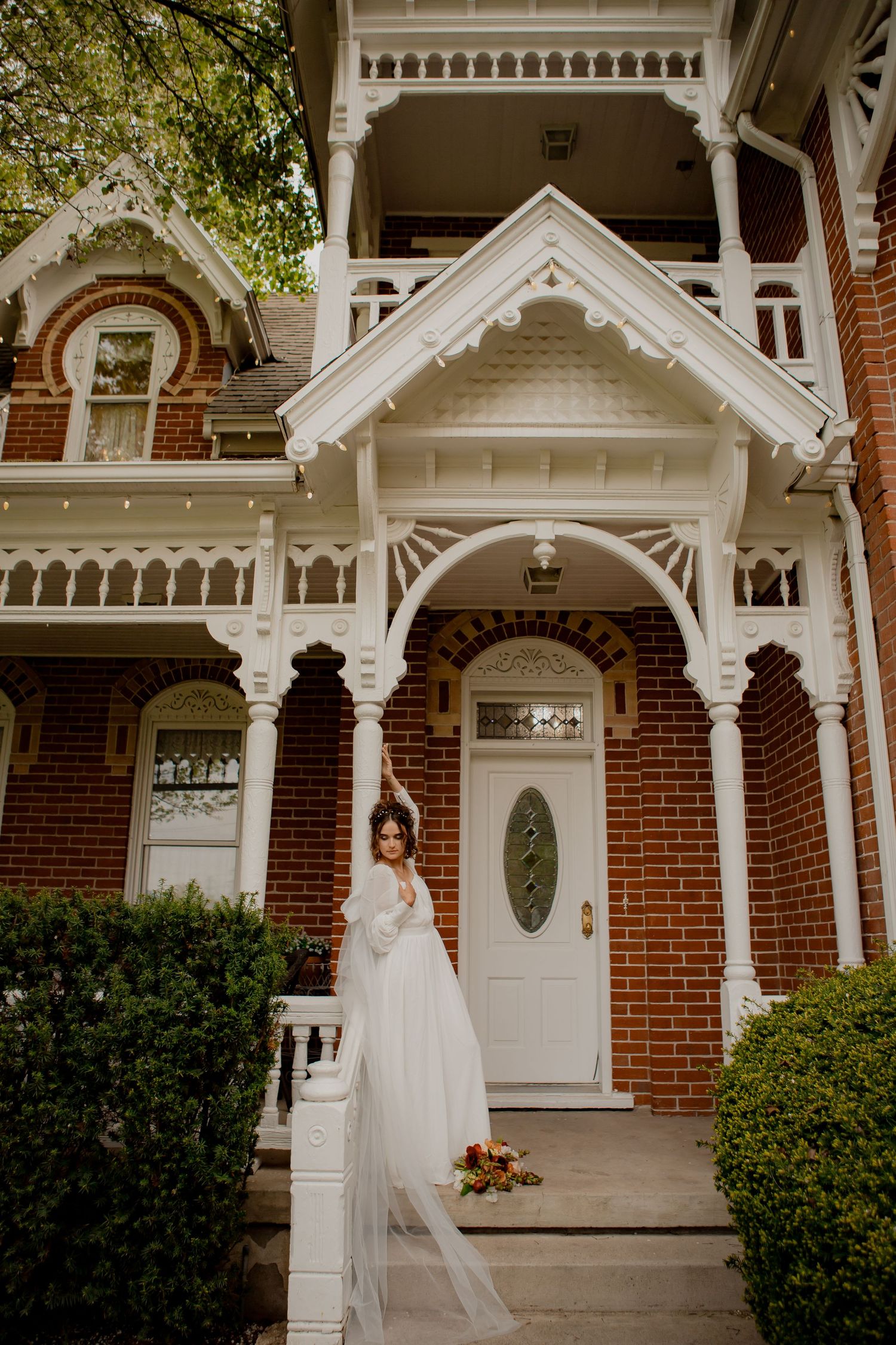 Bride in flowing gown stands gracefully on ornate Victorian porch with architectural details.