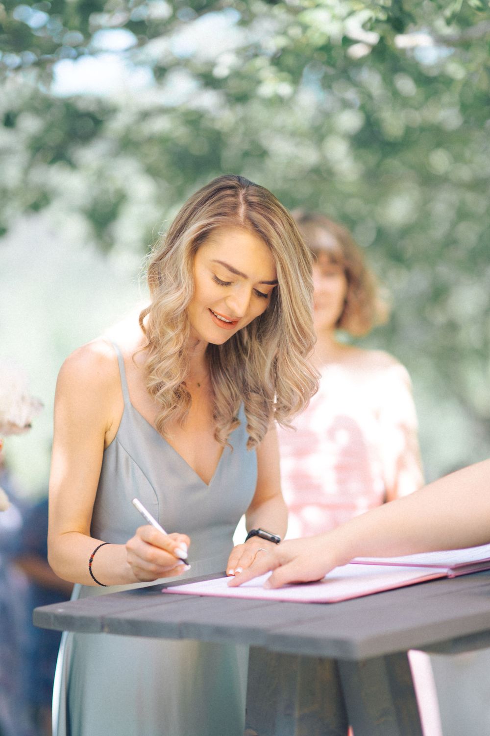 Woman in gray dress signs document at outdoor table with natural greenery backdrop.