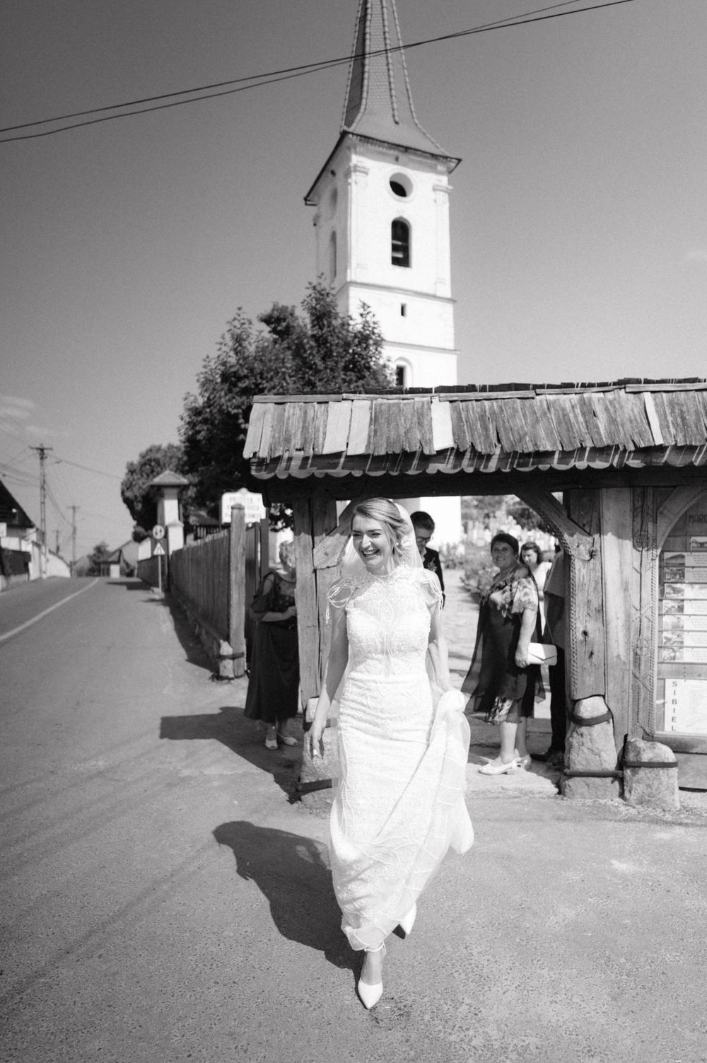 Wedding party walking down village street with white church steeple visible in background.