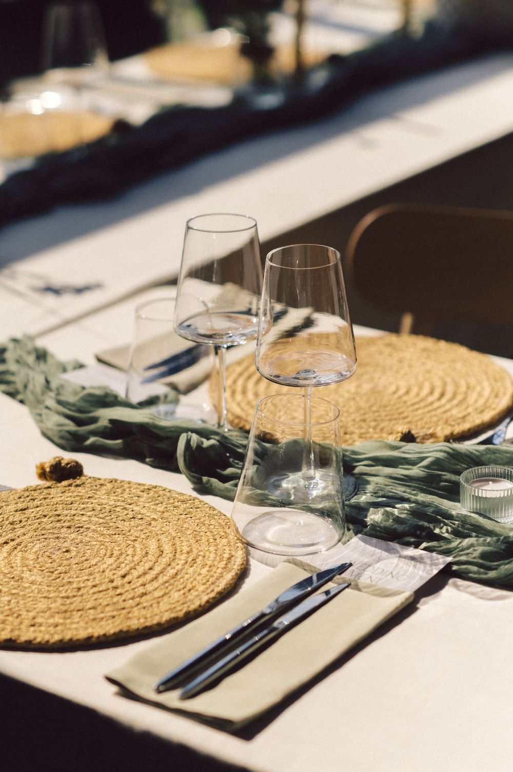 Place setting with woven placemat, wine glasses, and sage green fabric accent on white tablecloth.
