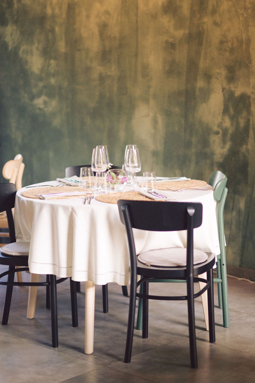 Round dining table with white tablecloth and black chairs against textured green wall.