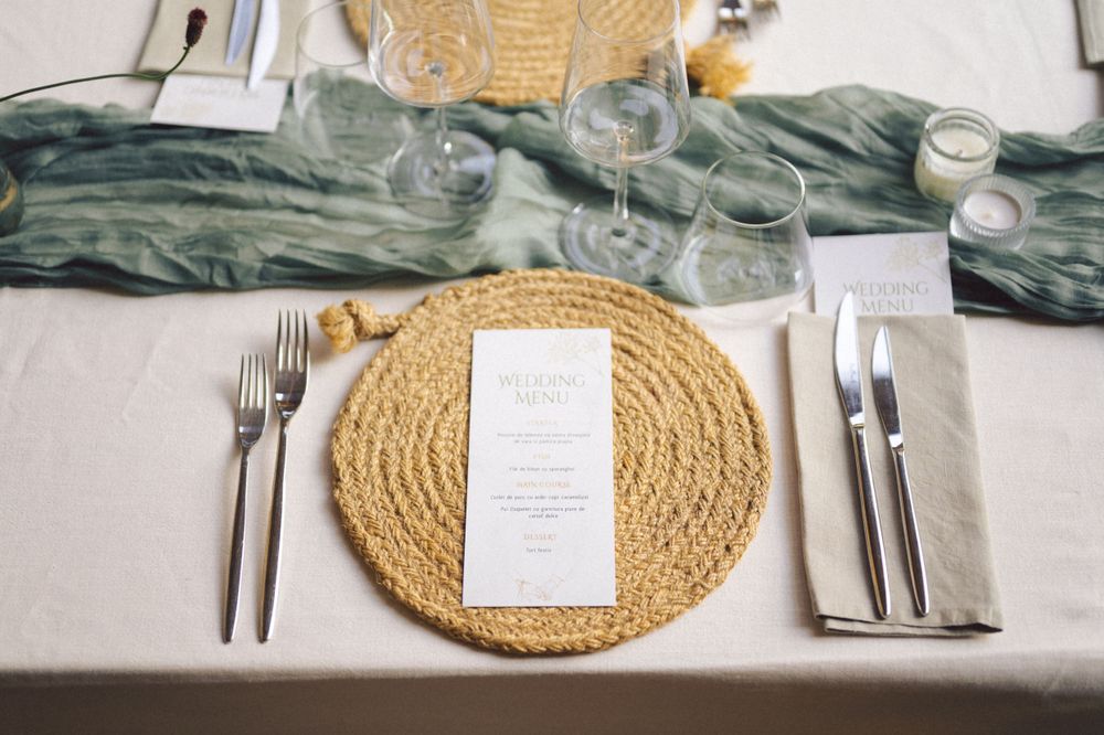 Wedding place setting with woven placemat, menu card, silverware and sage green fabric accent.