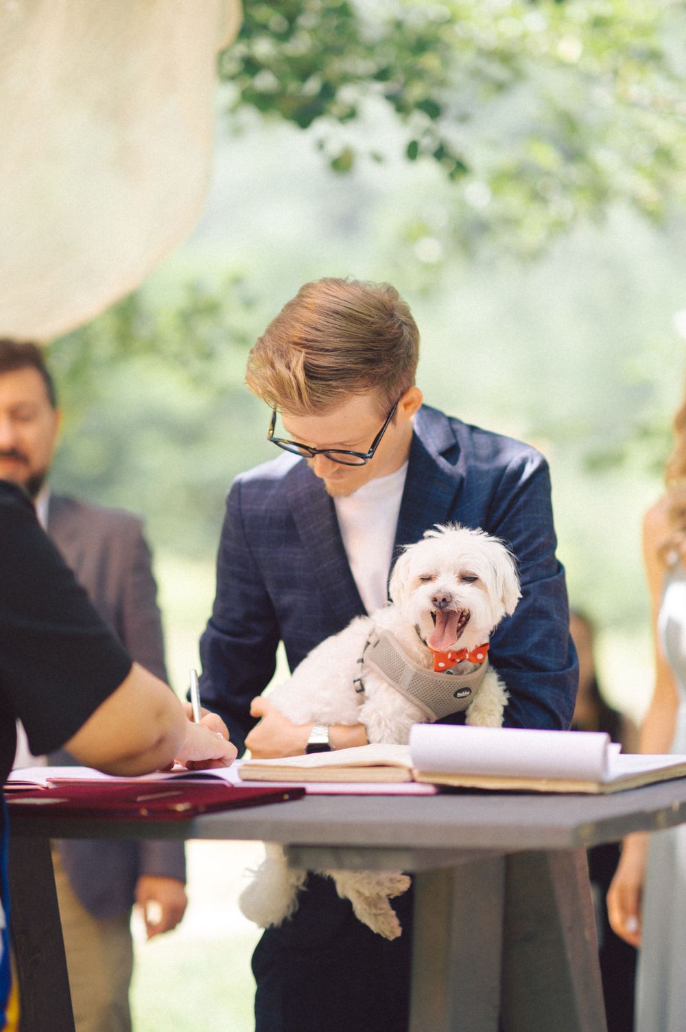 Person signs document while holding small white dog during outdoor ceremony.
