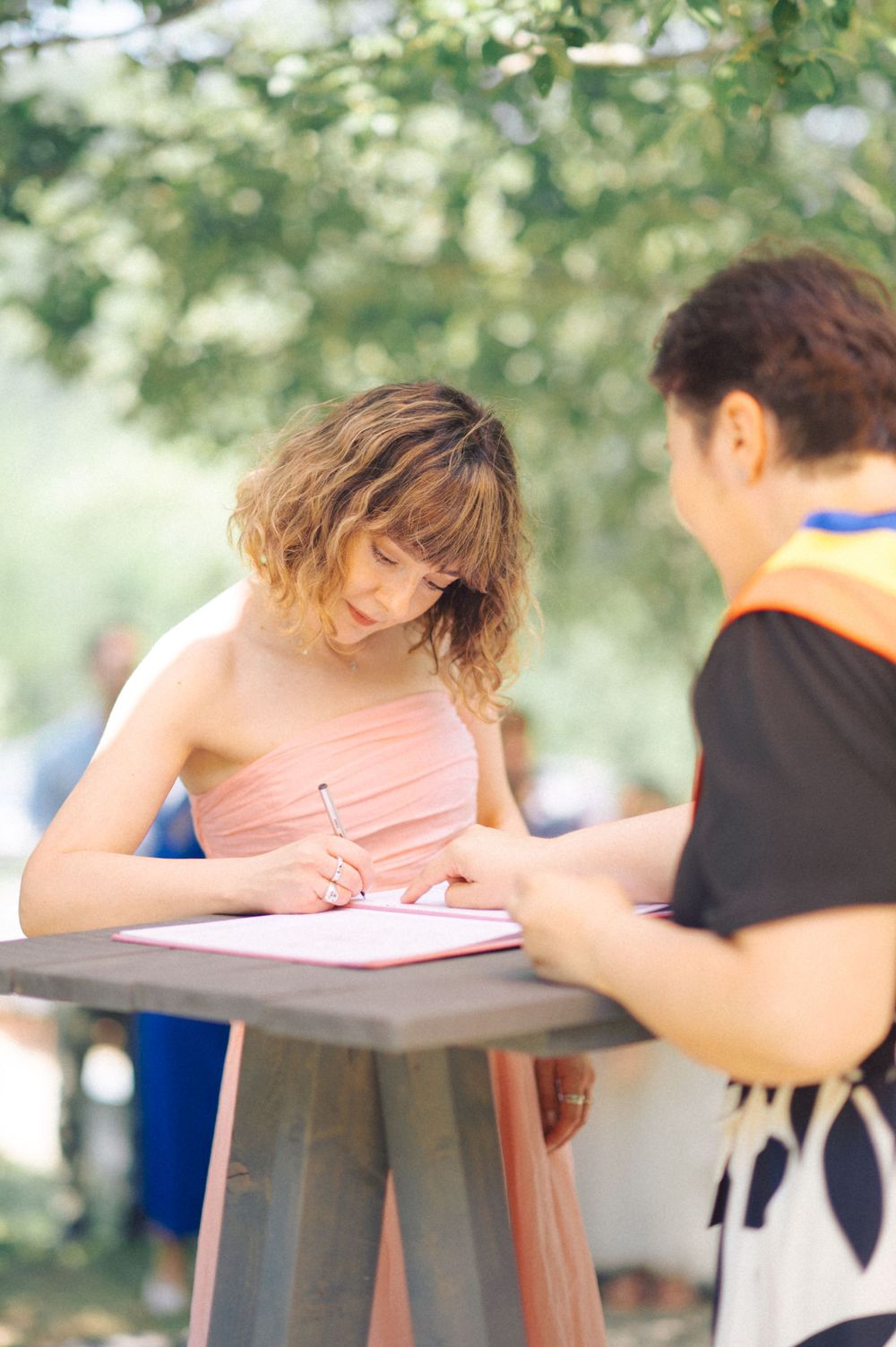 Guest in pink dress signs registry at outdoor ceremony table.