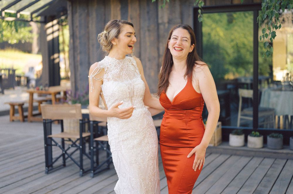 Two friends laugh together on a patio, one wearing a white lace dress and the other in a red satin gown.