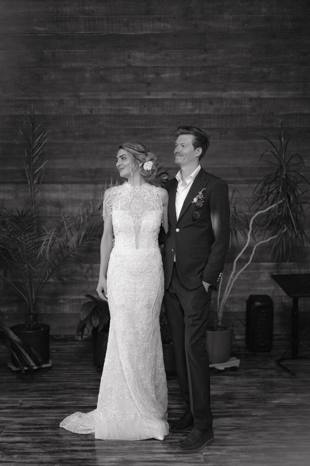 Black and white portrait of a couple in wedding attire against a wooden wall with plants.